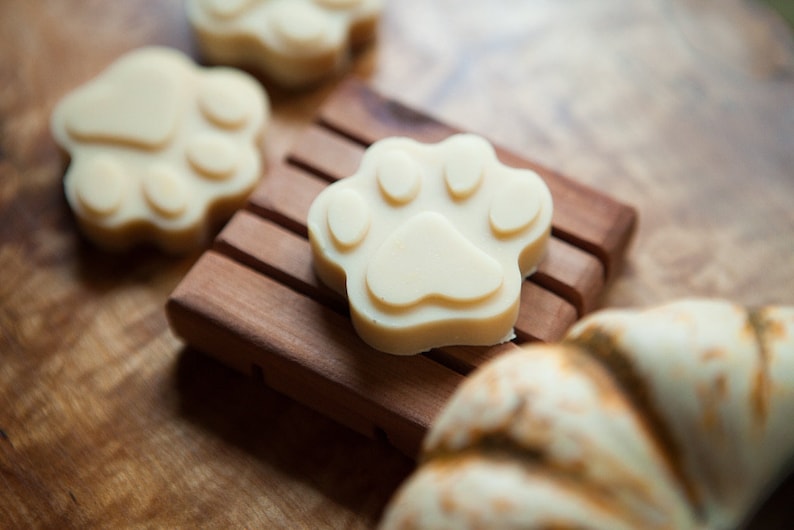 May include: Four white paw-shaped soaps on a wooden soap dish. The soaps are made in the shape of a dog's paw print.
