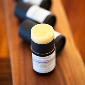 May include: A black and white label with the text "Happy Pet Skincare" and "Lavender Fresh" on a black tube with a yellow balm inside. The tube is sitting on a wooden surface.