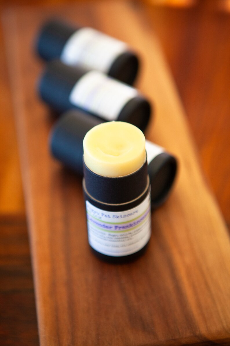 May include: A black and white label with the text "Happy Pet Skincare" and "Lavender Fresh" on a black tube with a yellow balm inside. The tube is sitting on a wooden surface.