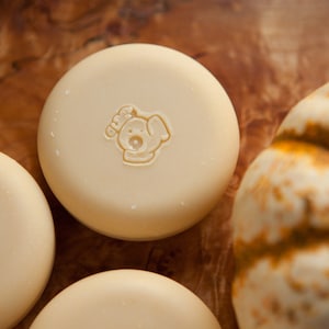 May include: Three round, white soap bars with a dog-shaped logo on each. The logo is brown and says "Woof!" on the top. The soap bars are on a wooden surface with a white pumpkin in the background.