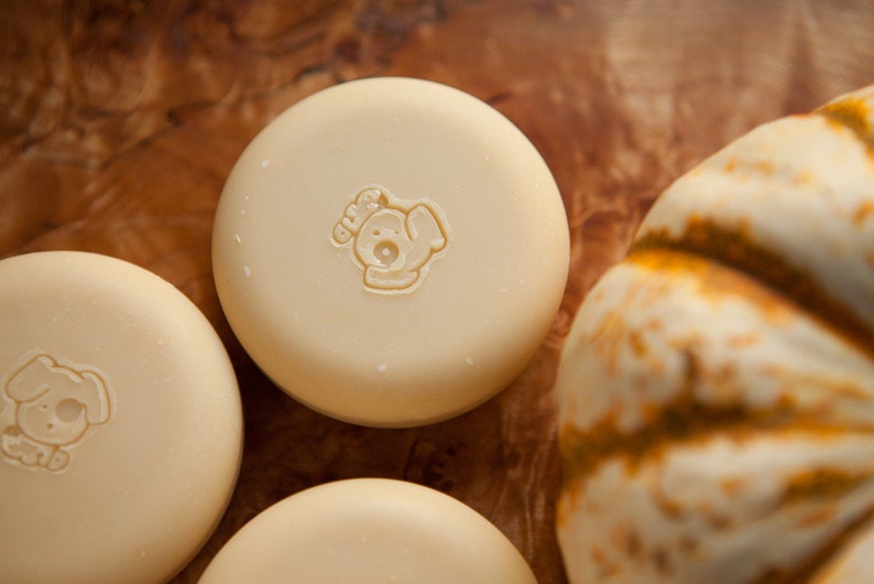May include: Three round, white soap bars with a dog-shaped logo on each. The logo is brown and says "Woof!" on the top. The soap bars are on a wooden surface with a white pumpkin in the background.