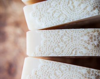 May include: Four bars of white soap with a delicate lace pattern pressed into the surface. The bars are stacked on top of each other.