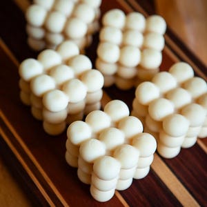 May include: Several white, cube-shaped candles with a bubble design are arranged on a wooden cutting board. The candles are made of a creamy white wax and have a smooth, rounded texture. The cutting board has a striped pattern.