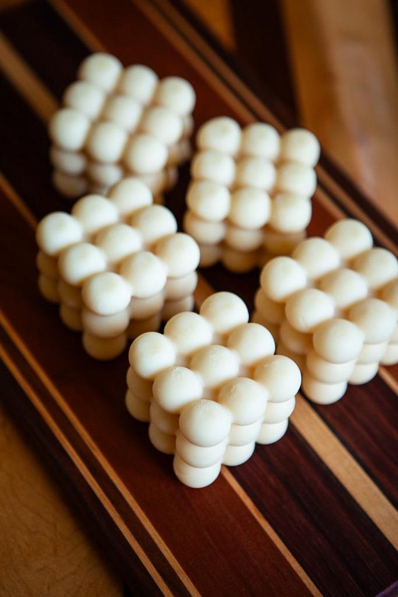 May include: Several white, cube-shaped candles with a bubble design are arranged on a wooden cutting board. The candles are made of a creamy white wax and have a smooth, rounded texture. The cutting board has a striped pattern.