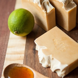 Puede incluir: Una cuchara de madera con miel, dos limas y cuatro barras de jabón hecho a mano. El jabón es marrón claro con un diseño de remolino blanco y tiene un patrón de panal de abeja en relieve en la parte superior.