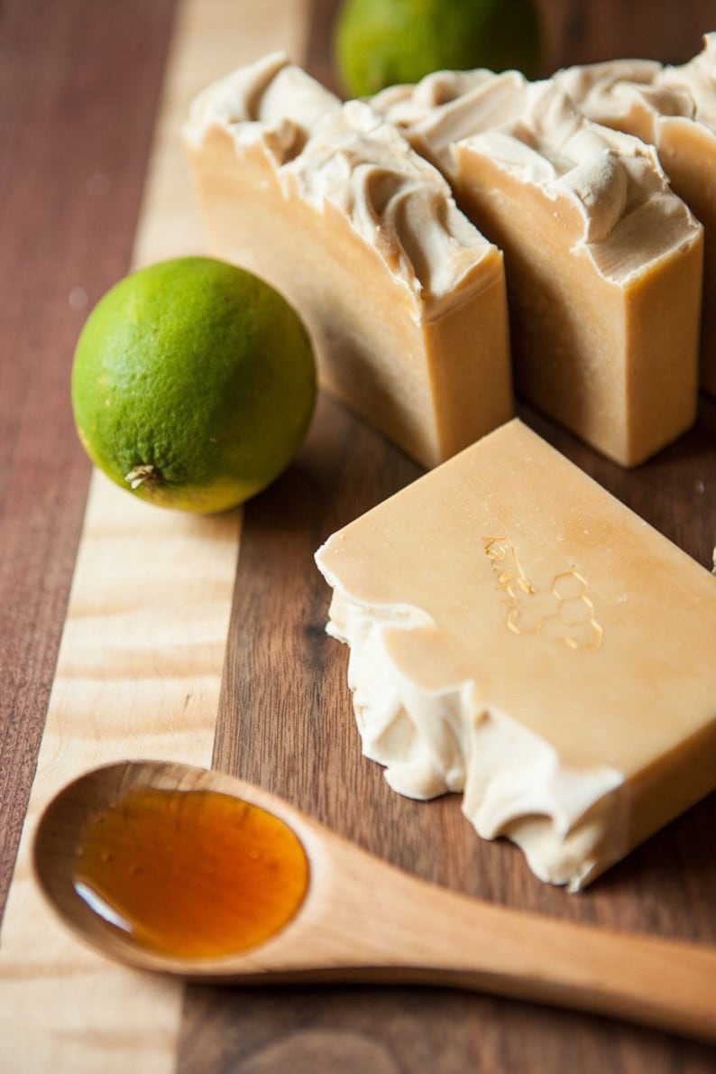 May include: A wooden spoon with honey, two limes, and four bars of handmade soap. The soap is light brown with a white swirl design and has a honeycomb pattern embossed on the top.