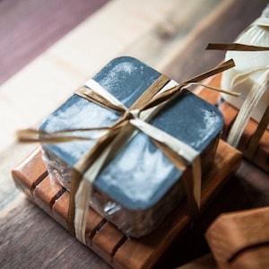 May include: Two bars of soap, one black and one white, are wrapped in brown twine and placed on wooden soap dishes.