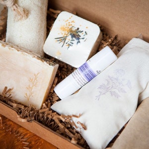 May include: A gift box filled with bath and body products. The box contains a bar of soap, a bath bomb, a lip balm, and a small pouch. The products are all white with lavender accents.
