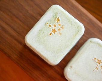 May include: Two rectangular bars of light green soap with rounded corners. Each bar features a stamped design of a cluster of small stars on the top surface, with the star indentations showing a slightly darker, golden-brown hue.