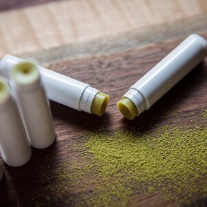 May include: Four white lip balm tubes with green balm inside, scattered on a wooden surface with green powder around them.