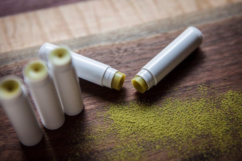 May include: Four white lip balm tubes with green balm inside, scattered on a wooden surface with green powder around them.