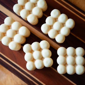 May include: Five off-white, bubble-shaped wax melts arranged on a wooden cutting board. The board has a striped pattern of dark and light brown wood. The melts are clustered in a grid-like pattern.