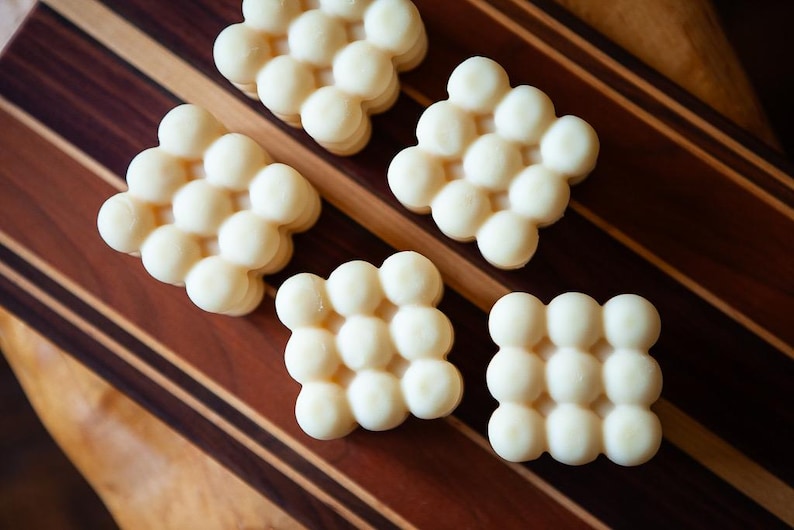 May include: Five off-white, bubble-shaped wax melts arranged on a wooden cutting board. The board has a striped pattern of dark and light brown wood. The melts are clustered in a grid-like pattern.