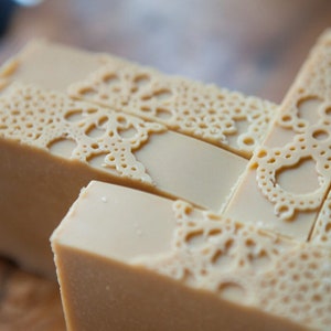 Pu&ograve; includere: Primo piano di una pila di saponette fatte a mano con un delicato motivo a pizzo impresso sulla superficie. Il sapone &egrave; di colore beige chiaro.