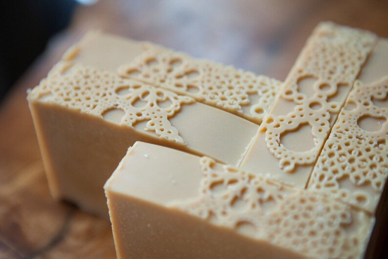 Pu&ograve; includere: Primo piano di una pila di saponette fatte a mano con un delicato motivo a pizzo impresso sulla superficie. Il sapone &egrave; di colore beige chiaro.