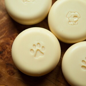 May include: Five round, white, unscented soap bars with different animal paw prints embossed on the top. The soap bars are arranged in a semi-circle on a wooden surface.