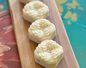 May include: Four off-white, hexagonal-shaped bars with intricate, embossed floral patterns on their top surfaces. These bars are arranged in a single, linear row along the center of a long, rectangular, natural-toned wooden serving board.