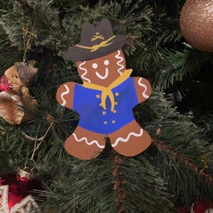 May include: A gingerbread man Christmas ornament with a blue uniform, a yellow scarf, and a black hat with a gold emblem. The ornament is hanging on a green Christmas tree.