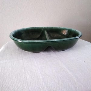 May include: A divided, oval-shaped serving bowl in a deep green hue. The ceramic bowl has a glossy finish and a light green rim. The design features two distinct compartments, ideal for serving snacks or dips. The bowl is placed on a white surface.