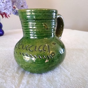 May include: A green ceramic pitcher with a handle and the word "Garcia" inscribed on the front. The pitcher has a bulbous base and a textured, ribbed design near the top. The background includes a blue vase and purple flowers.