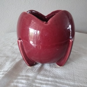 May include: A deep burgundy ceramic planter with a glossy finish, featuring a rounded body and three distinct, curved feet at the base. The top rim has a scalloped, heart-like shape with a wide opening, creating a smooth, continuous surface.