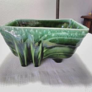 May include: A square, green ceramic planter with a glossy finish. The planter has a unique, textured design with three small feet. The color varies from light to dark green, creating a marbled effect. The planter is empty.