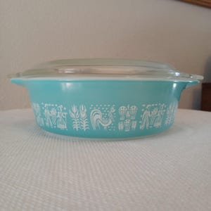 May include: A vintage turquoise Pyrex casserole dish with a clear glass lid. The dish features a white graphic design of a rooster, people, and wheat. The dish is round with handles on either side.
