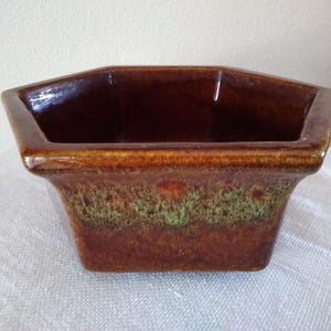 May include: A brown ceramic planter with a unique shape. The planter has a rectangular base and a flared, multi-sided top. The exterior features a band of green and brown speckled glaze, adding visual interest to the piece. The interior is a darker brown.