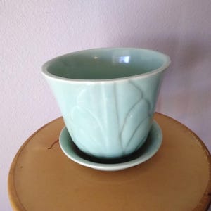 May include: A light green ceramic planter with a matching saucer. The planter has a flared shape and a leaf-like embossed design. The planter and saucer are sitting on a round, wooden table.