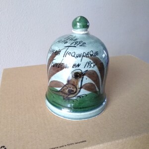 May include: A ceramic bell with a green and brown painted design. The bell has a small bird on the front and the text "Feliz Año 1992 Feria Tlaquepaque Fundada en 1955" on the side.