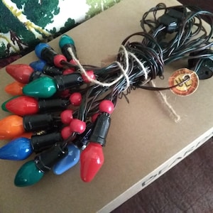 May include: A bundle of colorful Christmas lights with red, orange, green, and blue bulbs. The lights are connected by a black wire and tied with twine. The lights are on a cardboard box.