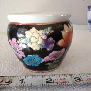 May include: A small, round ceramic pot with a black base and a white rim. The pot is decorated with colorful floral designs in pink, blue, yellow, and orange. A measuring tape shows the pot is approximately 3 inches wide.