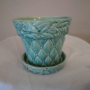 May include: A turquoise ceramic flower pot with a matching saucer. The pot features a diamond pattern and leaf detailing around the rim and saucer. The pot is designed to hold plants and flowers.