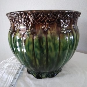 May include: A glazed ceramic planter featuring a deep green body with vertical fluting and a dark brown, intricate lattice-patterned rim. A white measuring tape is visible at the base, showing a measurement of approximately 8 inches in diameter.