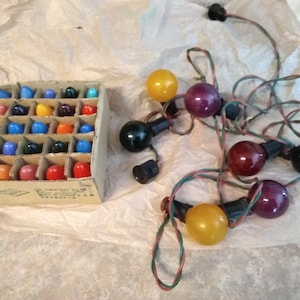 May include: A vintage set of Christmas lights with a variety of colorful bulbs in a cardboard box. The lights are in various colors, including red, blue, orange, and green. Some of the lights are strung together with a green and red wire.
