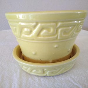 May include: A pale yellow ceramic planter with a matching saucer. The planter has a flared shape with a repeating geometric design around the top. The saucer also features the same design. The planter is designed to hold plants.