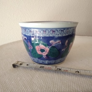 May include: A round, blue and white ceramic planter with a floral design. The planter has a white rim and a blue base. The floral design includes pink flowers and green leaves. The planter is approximately 4 inches in diameter.