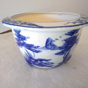 May include: A white ceramic planter with a blue and white landscape design. The planter has a wide, slightly flared rim with a blue border. The exterior features a detailed blue painted scene.