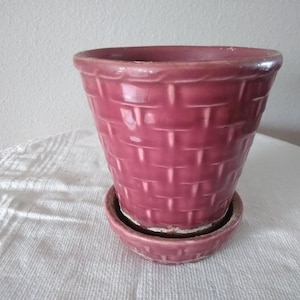 May include: A deep pink ceramic flower pot with a raised basket weave texture on the exterior. The pot features a matching circular saucer base with the same woven pattern, creating a cohesive look for this glazed, tapered plant container.