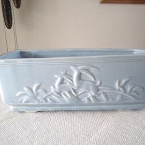 May include: A rectangular, light blue ceramic planter with a raised floral and deer design. The planter is approximately 12 inches long and 5 inches wide, with a slightly flared rim and small feet. Ideal for indoor or outdoor use.