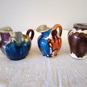 May include: Three handcrafted ceramic pitchers and a jar. The pitchers feature blue, orange, and brown glaze with handles. The jar is brown with a white base. These decorative pieces are suitable for home decor.