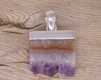 Puede incluir: Un colgante de plata con un cristal de amatista en bruto con matices púrpuras y blancas. El cristal está engastado en un bisel de plata y tiene un asa de plata para sujetarlo a una cadena.