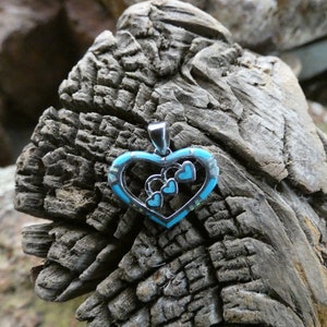 May include: A silver heart-shaped pendant with a turquoise inlay and smaller hearts inside. The pendant is on a rough wooden surface.