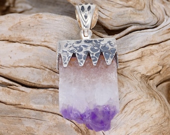 Puede incluir: Un colgante de plata con un cristal de amatista en bruto. El cristal es de color púrpura claro y tiene una superficie texturizada. El colgante está sobre un fondo de madera.