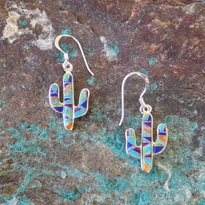 May include: A pair of silver earrings with a geometric cactus design. The cactus is made of turquoise, lapis lazuli, and other colorful stones.