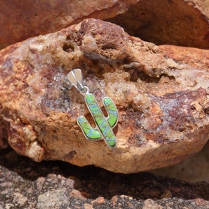 May include: A silver pendant shaped like a cactus with green and blue opal inlay. The pendant is sitting on a brown rock with white crystals.