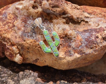 May include: A silver pendant shaped like a cactus with green and blue opal inlay. The pendant is sitting on a brown rock with white crystals.