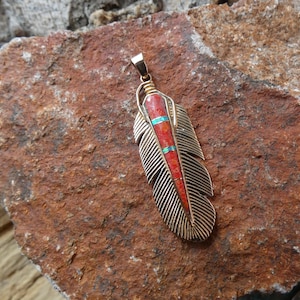 May include: A gold-coloured feather pendant with a central inlay of red and turquoise stones. The pendant is set against a textured, reddish-brown stone surface, showcasing the intricate details of the jewellery.