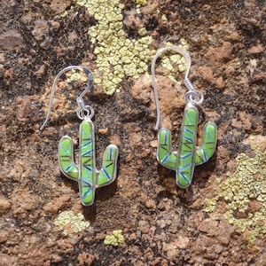 May include: A pair of sterling silver cactus earrings. Each earring features a green mosaic design with blue and silver accents. The earrings are set against a textured brown and tan background.