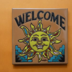 May include: Square ceramic tile with the word "WELCOME" in black letters above a smiling sun design. The sun is yellow with blue, red, and orange accents. The tile has a brown background and blue and black decorative elements.